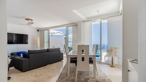 Una sala de estar moderna con sofá, mesa de comedor y sillas. Hay un televisor montado en la pared, y un balcón con vistas.