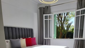 Ein Schlafzimmer mit einem großen Bett, grauen Vorhängen, einem Fenster mit Blick ins Grüne und einer modernen Deckenleuchte.