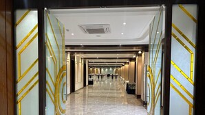 Corredor de hotel com piso de mármore polido, portas de vidro com detalhes dourados e uma fileira de mesas com cadeiras ao fundo.
