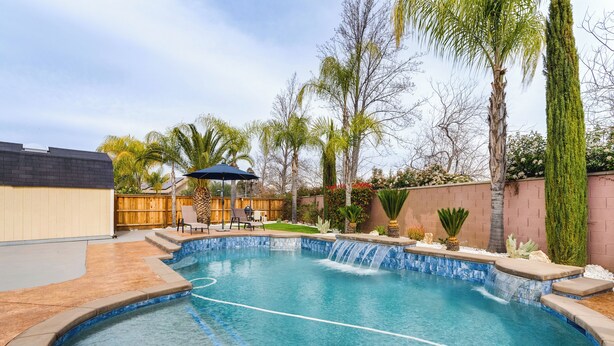 A swimming pool with a waterfall feature, surrounded by palm trees and a wooden fence.