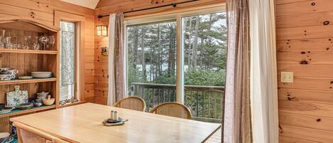 A wooden cabin interior with a dining table, chairs, a shelf with glassware, and a large window with curtains.