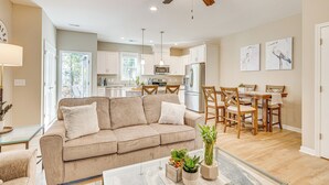 A spacious living room with a beige sofa, a dining area with wooden chairs, a ceiling fan, and a kitchen in the background. - Coastal Beaufort Abode - Pool Access, Near Beaches (Beaufort)
