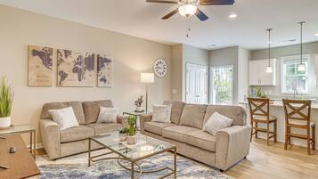 A modern living room with two sofas, a coffee table, and a dining area with chairs. There is a ceiling fan, a clock, and a world map on the wall.