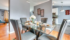 A modern dining area with a glass table, grey upholstered chairs, and a kitchen in the background.