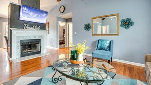 A living room with a fireplace, a television mounted above it, a blue armchair, and a glass coffee table with decorative items.