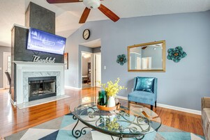 A living room with a fireplace, a television mounted above it, a blue armchair, and a glass coffee table with decorative items.