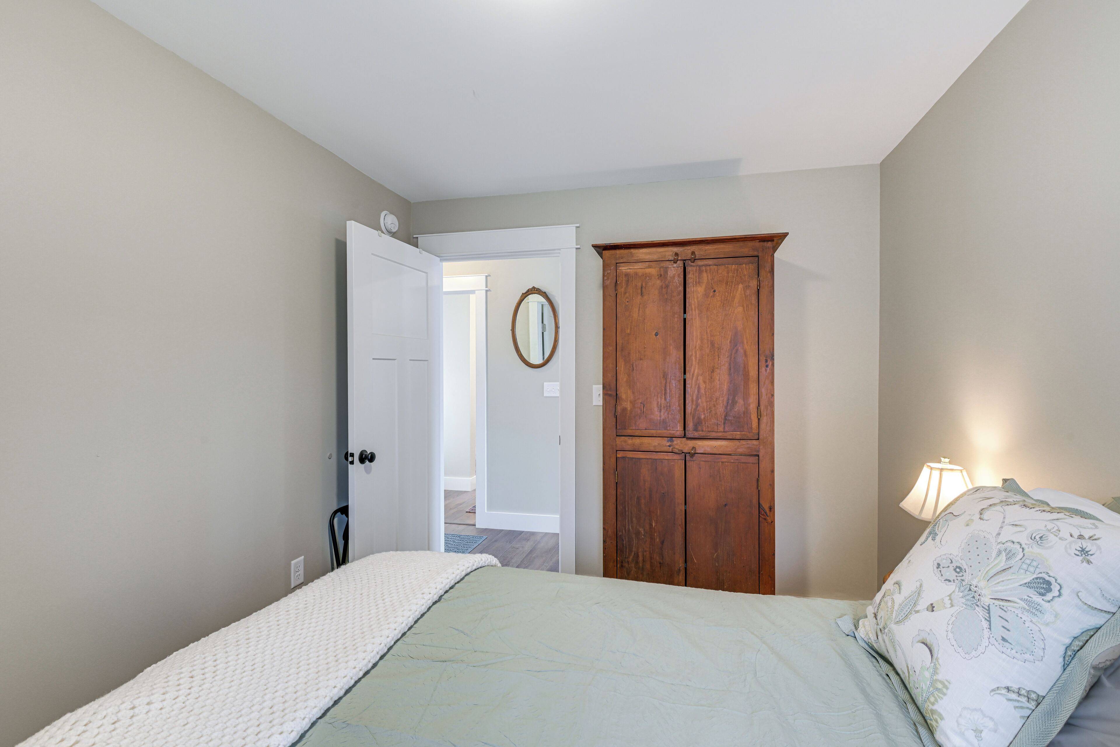 Un dormitorio con una cama, un armario de madera, una puerta que da a otra habitación y una lámpara de noche.