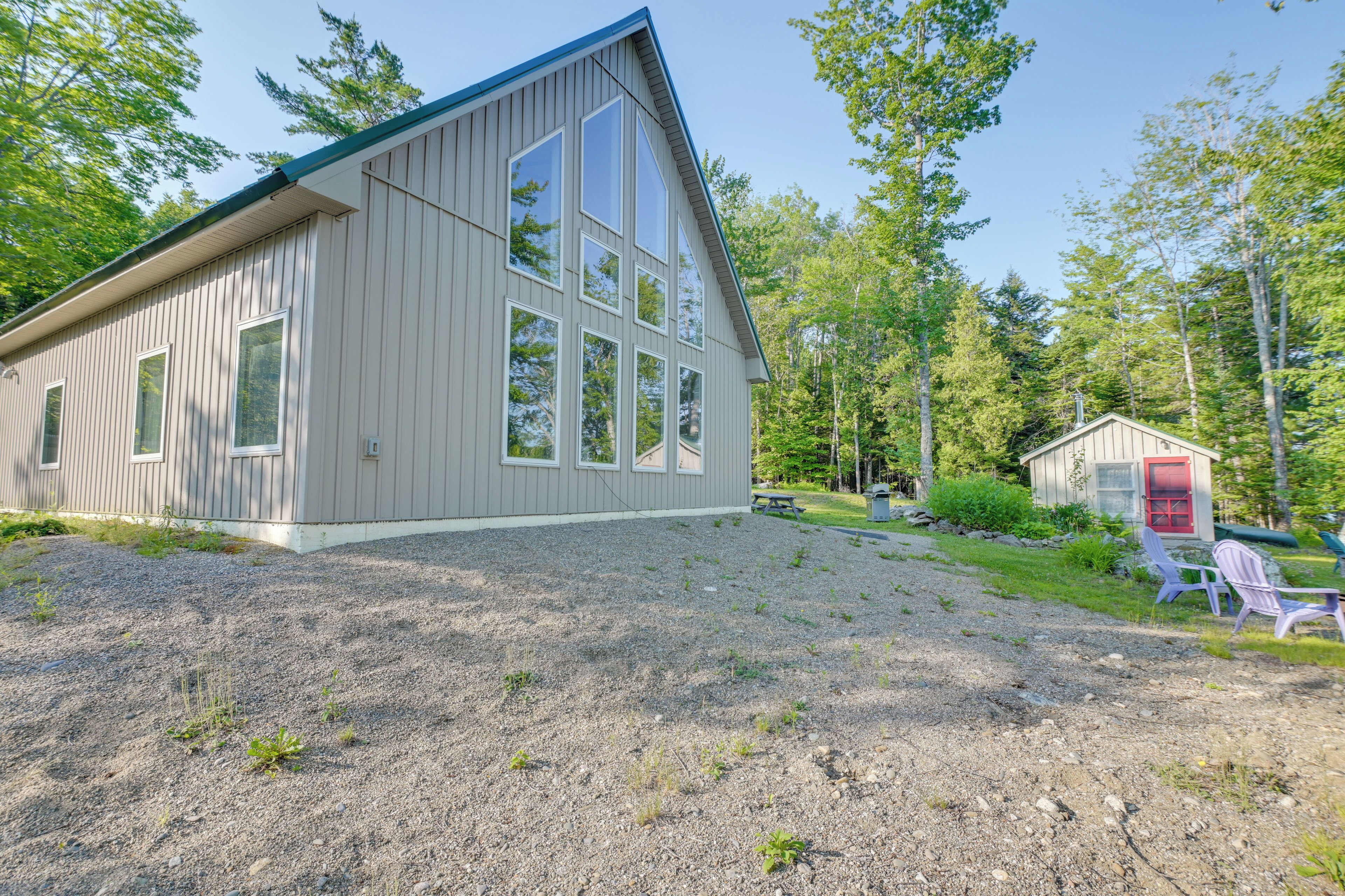 A wooden cabin with large windows, a gravel driveway, and a small shed in a wooded area.