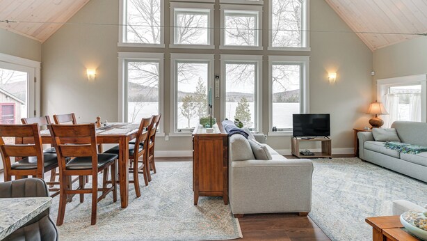 A spacious living area with a high ceiling, large windows, a dining table with chairs, a sofa, a TV, and a lamp. - Lakefront Bucksport Home w/ Fire Pit + Gas Grill! (Bucksport)