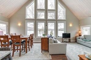 A spacious living area with a high ceiling, large windows, a dining table with chairs, a sofa, a TV, and a lamp. - Lakefront Bucksport Home w/ Fire Pit + Gas Grill! (Bucksport)