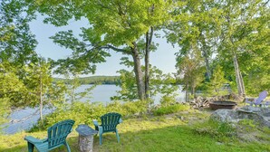 A lakeside setting with two Adirondack chairs, a fire pit, and a stone stump. - Lakefront Bucksport Home w/ Fire Pit + Gas Grill! (Bucksport)
