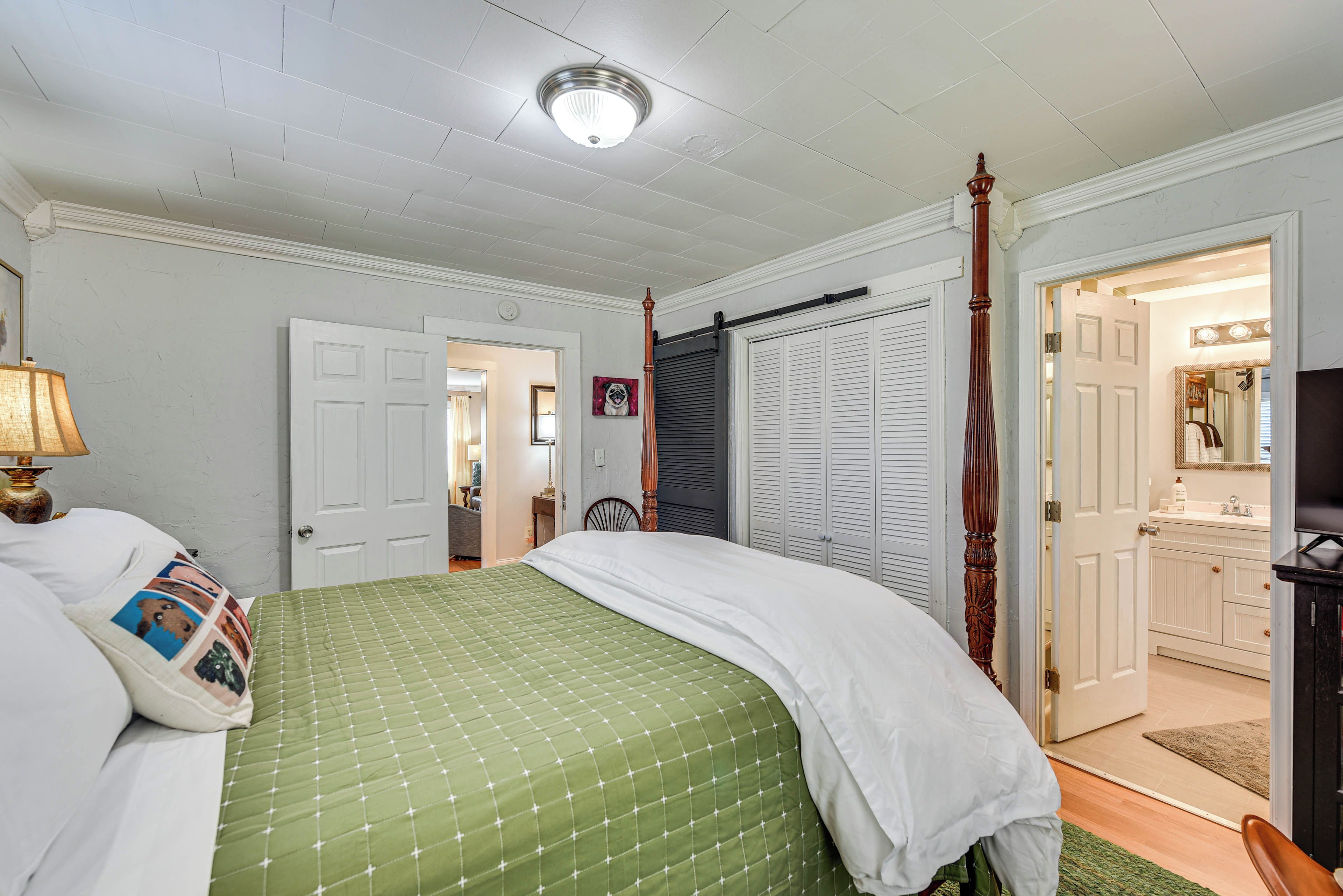 Un dormitorio con edredón verde acolchado, ropa de cama blanca, una cama con dosel de madera, una mesita de noche con lámpara y una puerta que da a un baño.