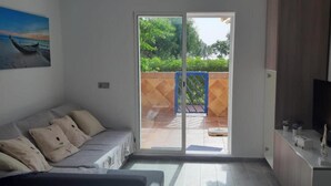A modern living room with a sofa, a television, and a sliding glass door leading to an outdoor area with a blue gate and greenery. - Studio PREMIUM Vera Playa Naturist  (Vera)