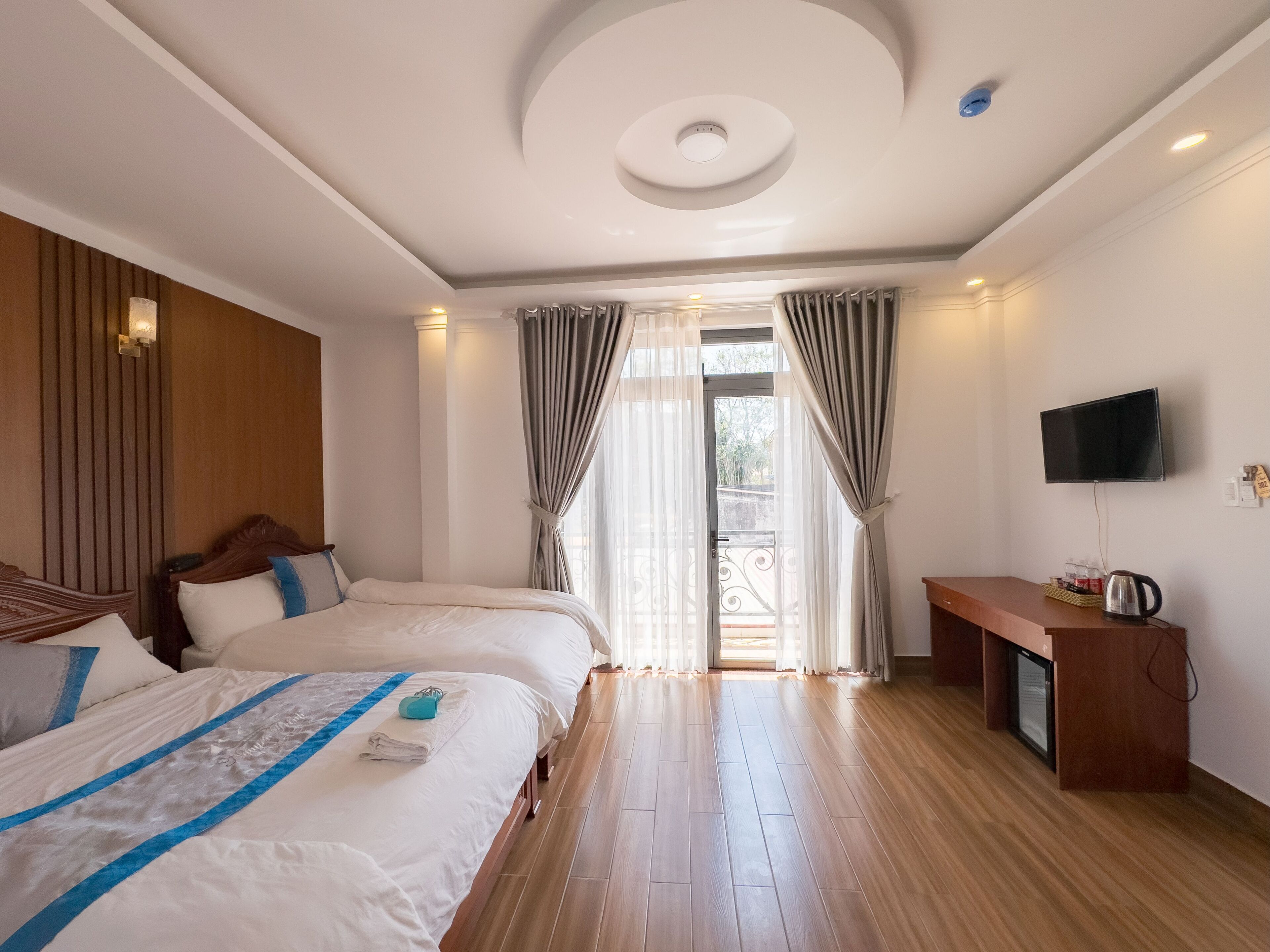 A hotel room with two beds, a wooden floor, a TV mounted on the wall, and a desk with a kettle.
