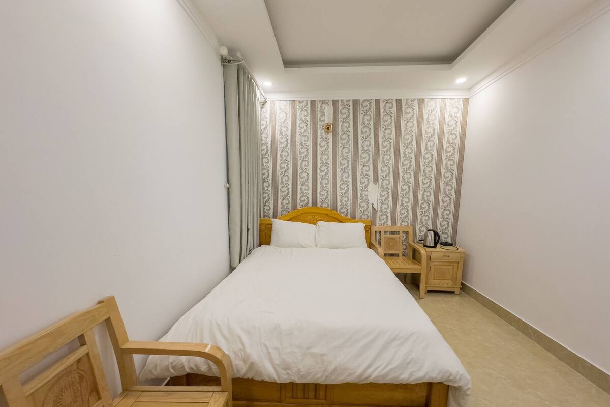 A bedroom with a wooden bed, a wooden chair, a nightstand with a kettle, and patterned wallpaper.