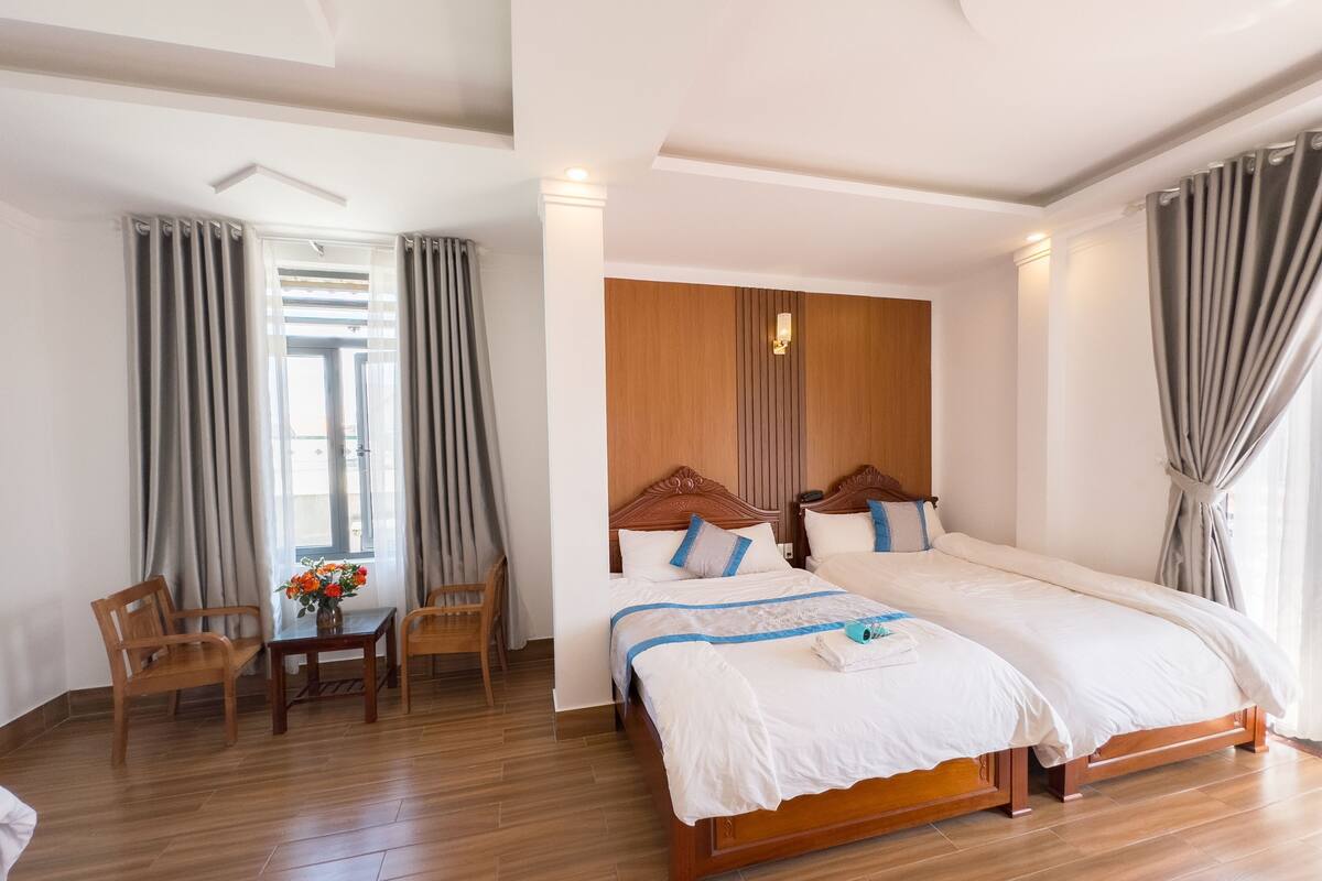 A hotel room with two beds, wooden flooring, a wooden headboard, a small table with a vase of flowers, and a window with curtains.