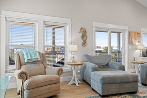 A living room with two armchairs, a sofa, a small table, and a lamp. There are large windows with blinds, a seahorse wall decoration, and a Florida map on the wall.