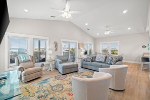 A spacious living room with a high ceiling, large windows, and a ceiling fan.