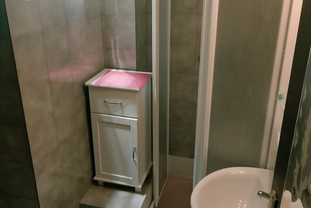 Een badkamer met een wit wastafelmeubel, een klein kastje met een roze bovenblad, en een douchecabine van glas.