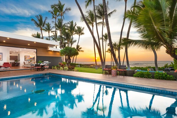 A modern pool area with a view of the sunset and ocean, surrounded by palm trees. - Malulani Villa, Luxury Oceanfront 3 BR, Private Pool, Hot Tub & Pickleball! (Kamuela)