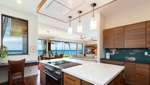 A modern kitchen with a large island, wooden cabinets, and a view of the ocean. - Malulani Villa, Luxury Oceanfront 3 BR, Private Pool, Hot Tub & Pickleball! (Kamuela)