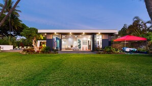 A modern house with a well-maintained lawn, outdoor seating area, and a red umbrella. - Malulani Villa, Luxury Oceanfront 3 BR, Private Pool, Hot Tub & Pickleball! (Kamuela)