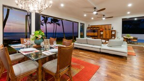 A modern living room with a large glass table set for dining, a sofa, and a ceiling fan. - Malulani Villa, Luxury Oceanfront 3 BR, Private Pool, Hot Tub & Pickleball! (Kamuela)