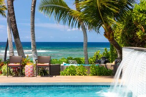 A poolside area with chairs, a small table, and a waterfall feature. - Malulani Villa, Luxury Oceanfront 3 BR, Private Pool, Hot Tub & Pickleball! (Kamuela)