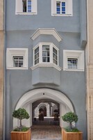 Un edificio con fachada gris, detalles blancos y tres ventanas con marcos blancos. Dos macetas de madera con plantas se encuentran frente a la entrada.