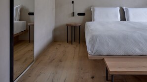 Un dormitorio minimalista con un espejo grande, una cama con ropa de cama blanca, una mesita de noche y un banco de madera.