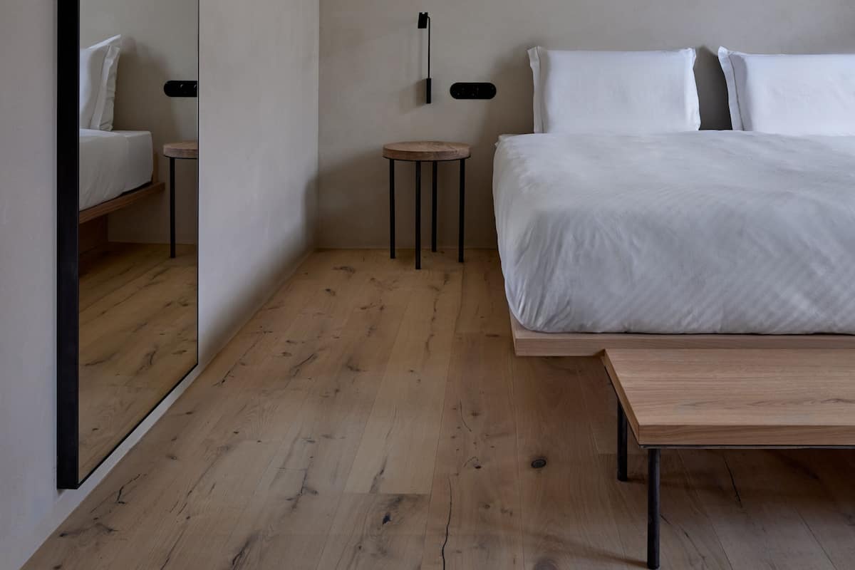 A minimalist bedroom with a large mirror, a bed with white linens, a bedside table, and a wooden bench.