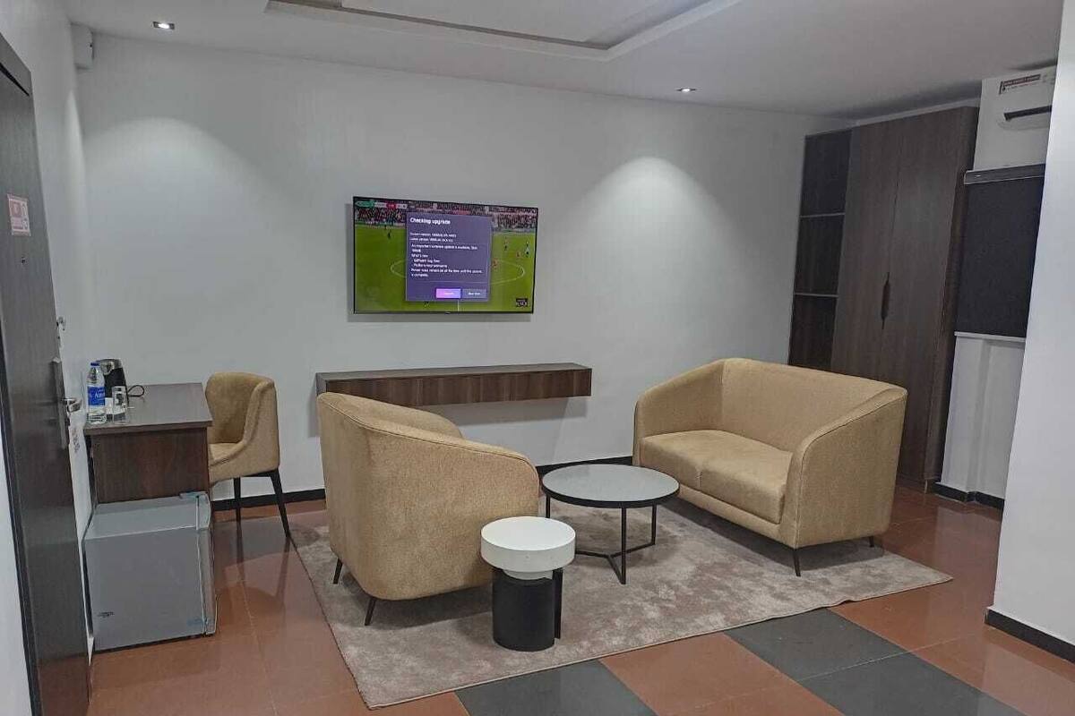 A modern office space with two beige armchairs, a small round table, a desk with a computer, and a flat-screen TV mounted on the wall.