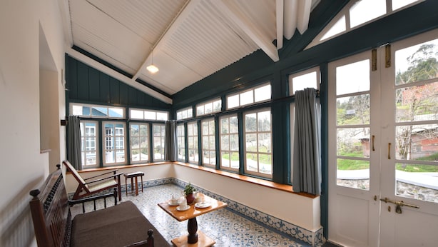 A room with a large bay window, a wooden bench, a small table with a plant, and a tiled floor. - The Hearth (Kodaikanal)