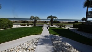 Caminho que leva a uma praia, com vista clara para o oceano, cercado por vegetação cuidada e um edifício à direita.
