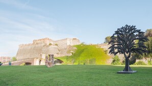 Un área con césped, una gran estructura de piedra al fondo y una escultura de metal que parece un árbol.