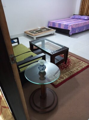 A small room with a glass-top table, a sofa, a bed with a purple bedspread, and a patterned rug.