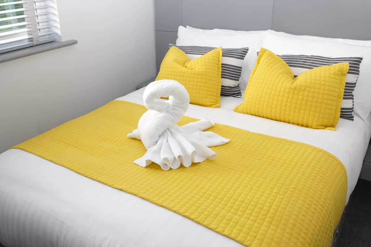 A neatly made bed with a yellow quilt, white pillows, and a white towel arranged in a swan shape.
