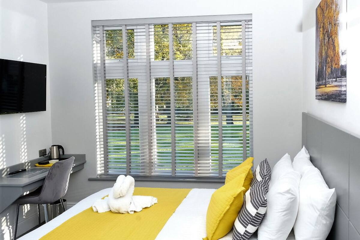 A hotel room with a bed, a desk, a chair, a flat-screen TV, and a window with blinds.