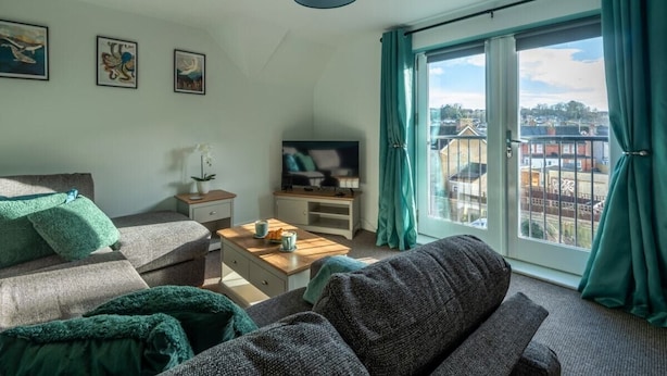 A living room with a sofa, a coffee table, and a TV. There are curtains on the window, and artwork on the wall.