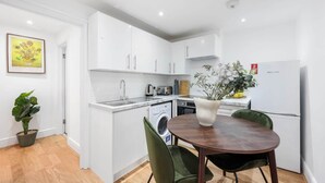 A compact kitchen with white cabinets, a sink, a washing machine, and a dining area with a table and chairs. - Classy&Stylish 2 bed flat (London)