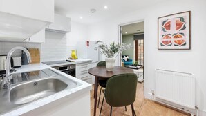 A modern kitchen with white cabinetry, a stainless steel sink, and a dining area with a table and chairs. - Classy&Stylish 2 bed flat (London)
