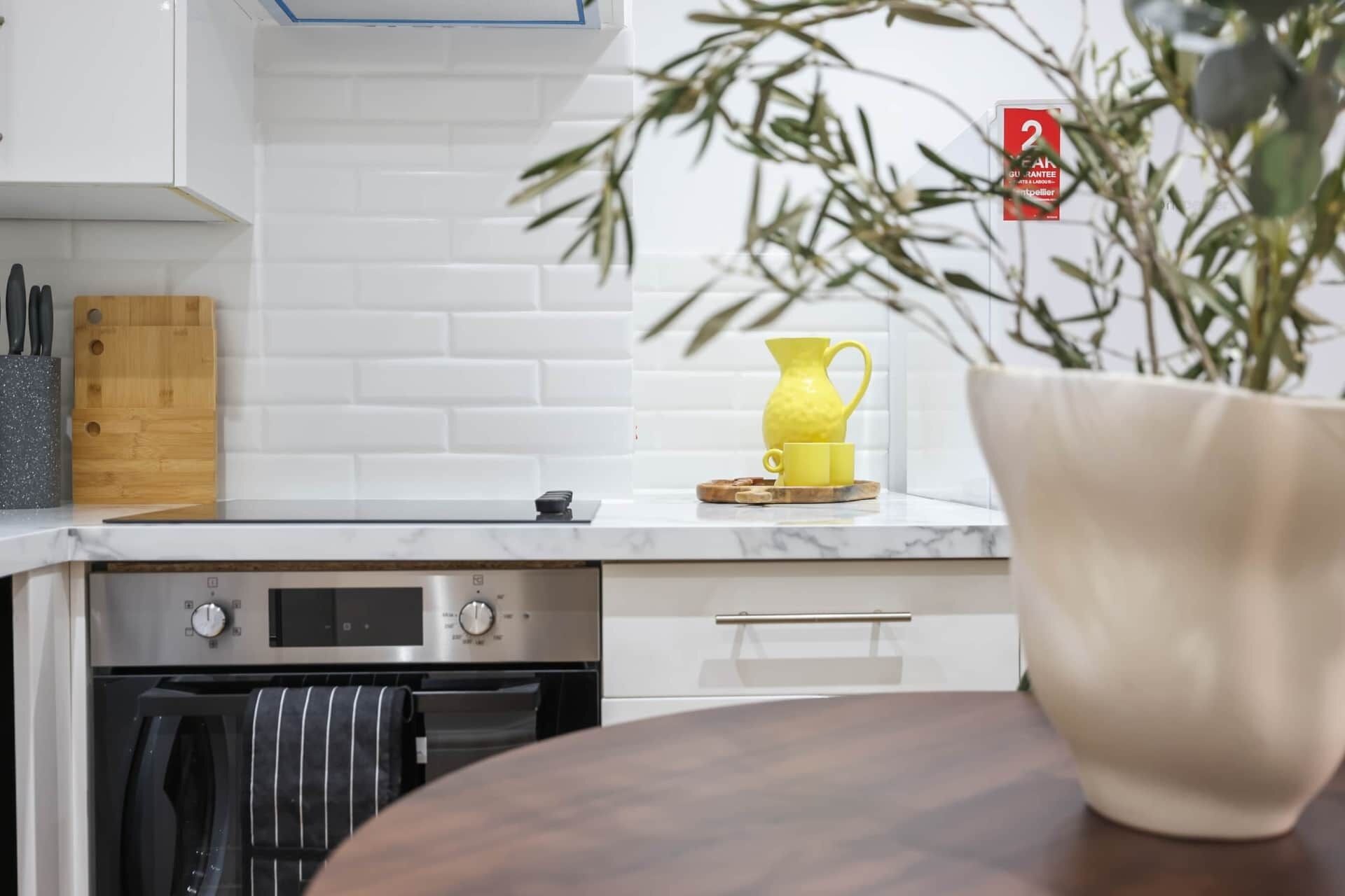 Een keuken met witte kastjes, een vaatwasser, een oven en een aanrecht met een gele kan en beker.