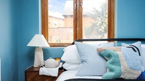 A bedroom with a bed, a nightstand with a lamp, a teapot, and a window with a view of buildings. - Classy&Stylish 2 bed flat (London)