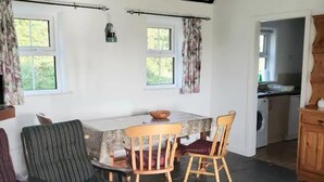 A dining area with a table and chairs, a washing machine in the background, and a view of the outdoors through windows. - Traditional Holiday Cottage Louisburgh. (Louisburgh)