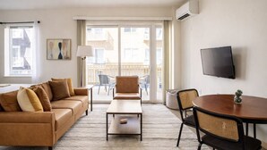En moderne stue med sofa, sofabord og spisebord, et tv ophængt på væggen, samt en balkon med udendørs møbler.