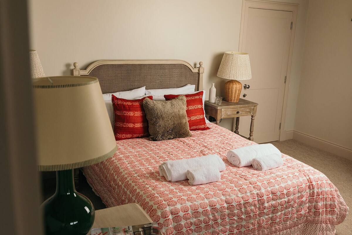 A neatly made bed with a patterned bedspread, two pillows, and two rolled towels. There is a bedside table with a lamp and a small decorative item.