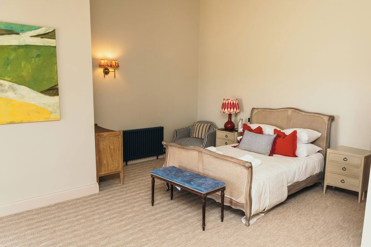 A bedroom with a bed, bedside table, lamp, bench, and a painting on the wall.