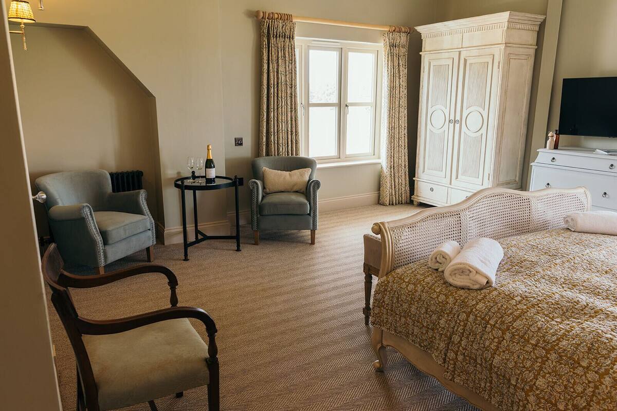A hotel room with a bed, two armchairs, a small table with a bottle and glasses, a wardrobe, and a television.