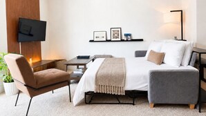 Une chambre d’hôtel moderne équipée d’un lit, d’un canapé, d’un fauteuil, d’une télévision, d’une petite table et d’une plante.