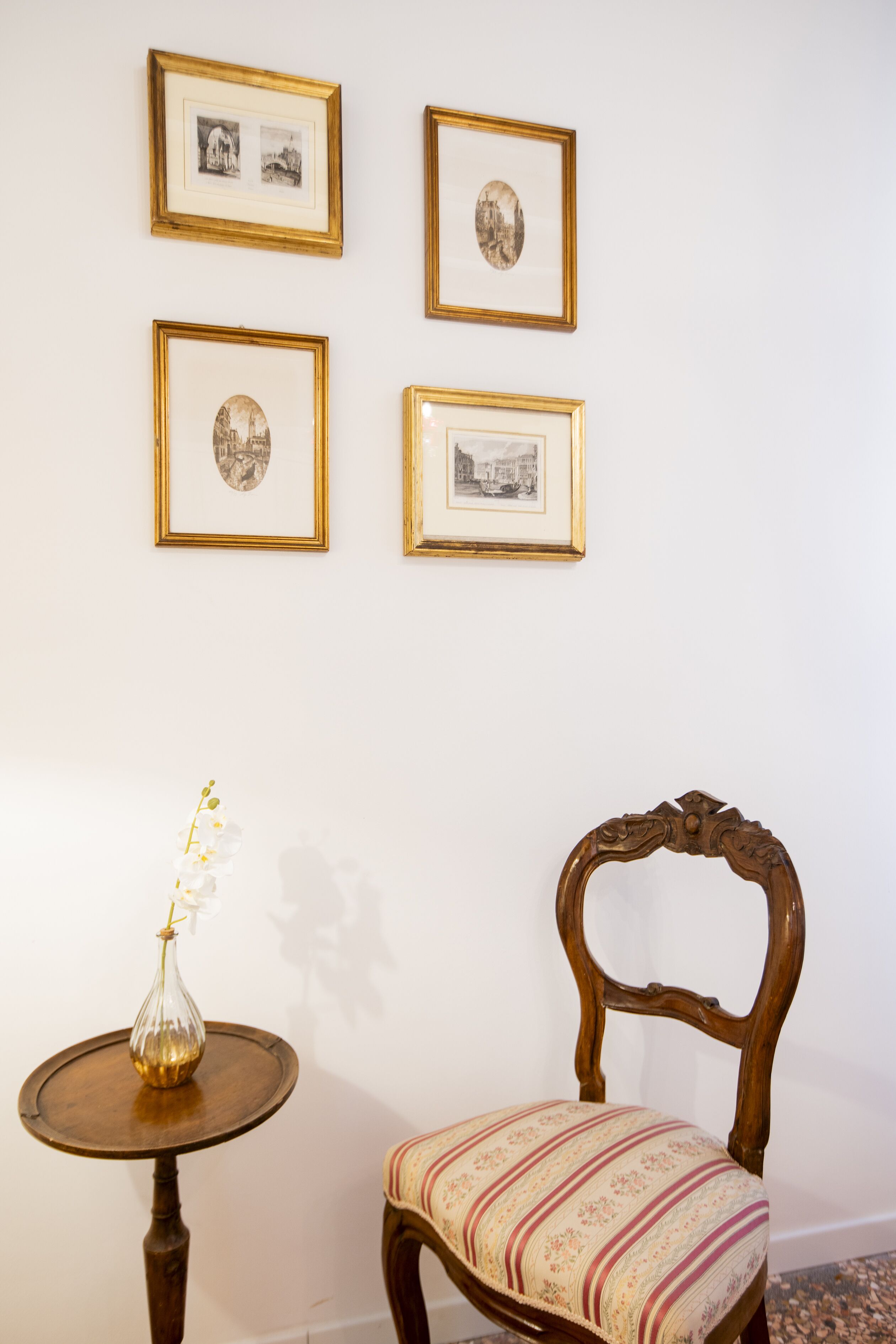 Una habitación con una silla de madera y una mesita con un florero. Cuatro cuadros enmarcados decoran la pared.